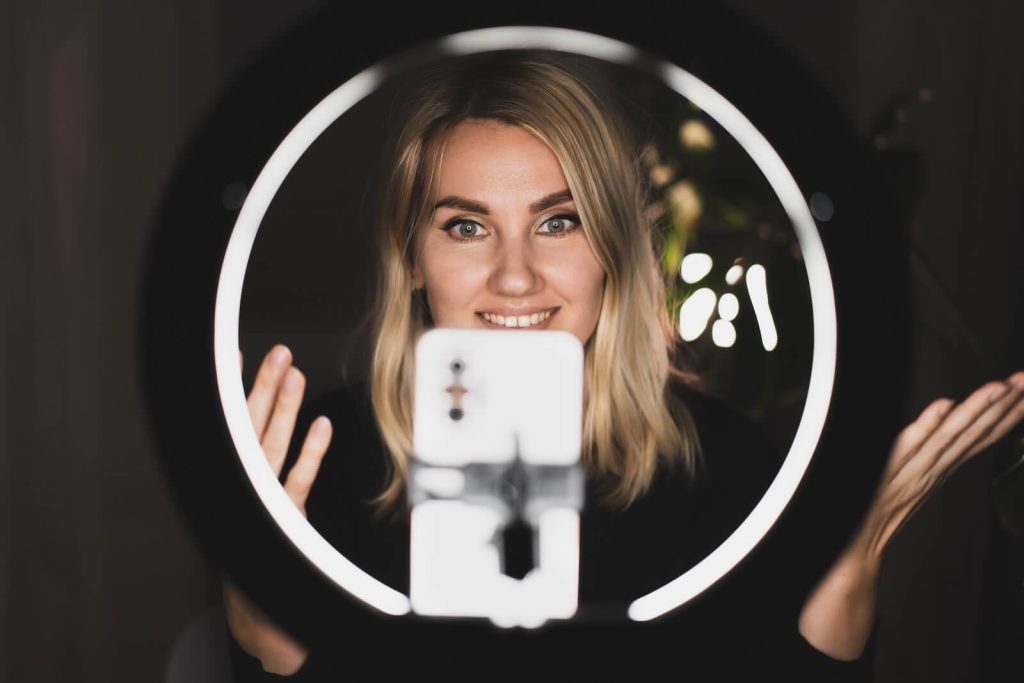 A blonde woman shooting a video of herself with a ring light setup.