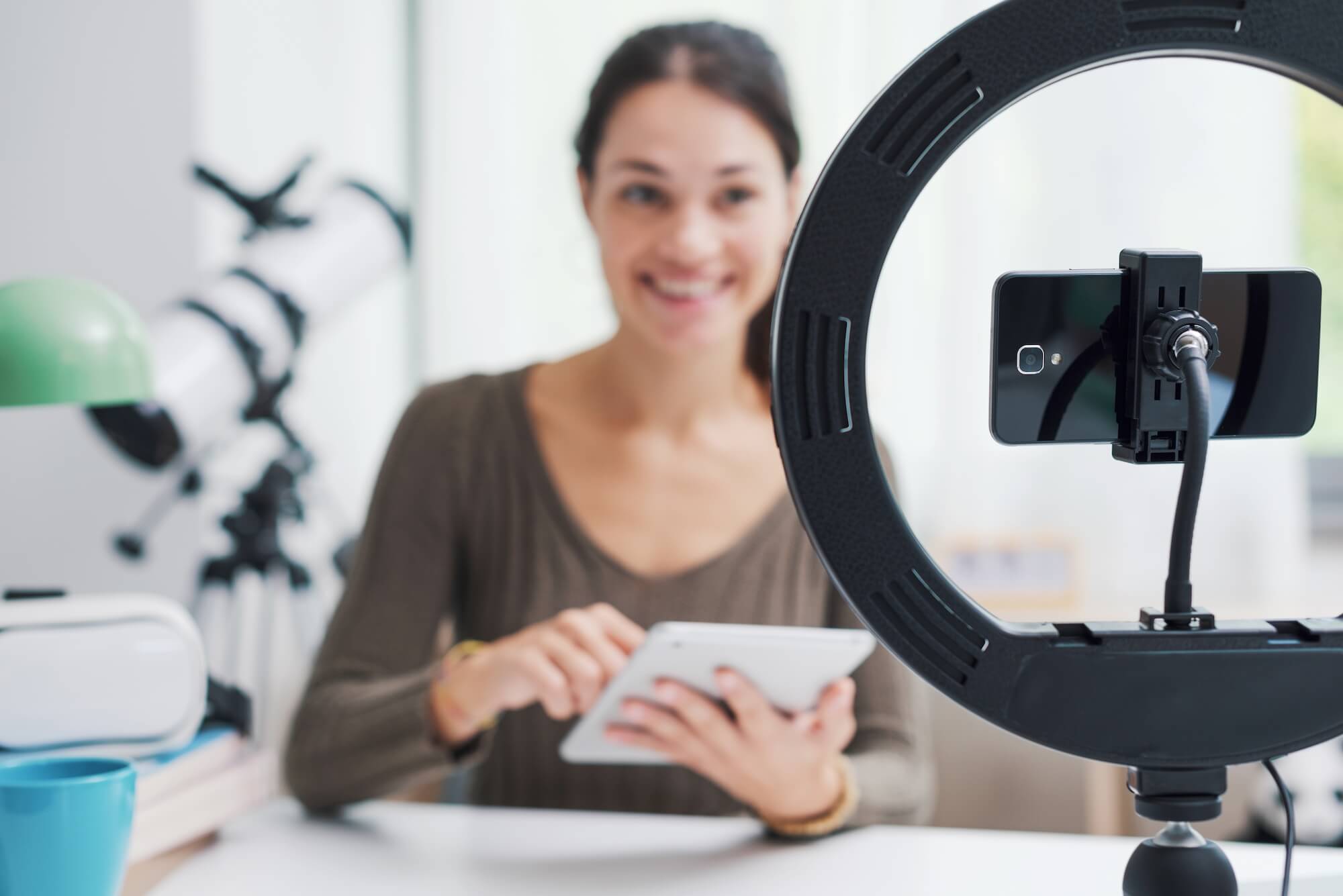 A woman is filming on her phone with a ring light while using her tablet.