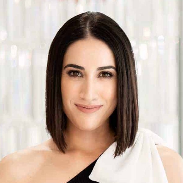 A headshot of a woman with shoulder-length hair is wearing a black top with a big white bow on the shoulder.