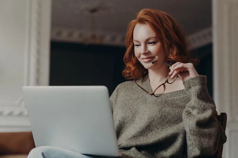 A woman wearing a sweater with red hair looks happy while staring at her laptop, holding her eyeglasses.