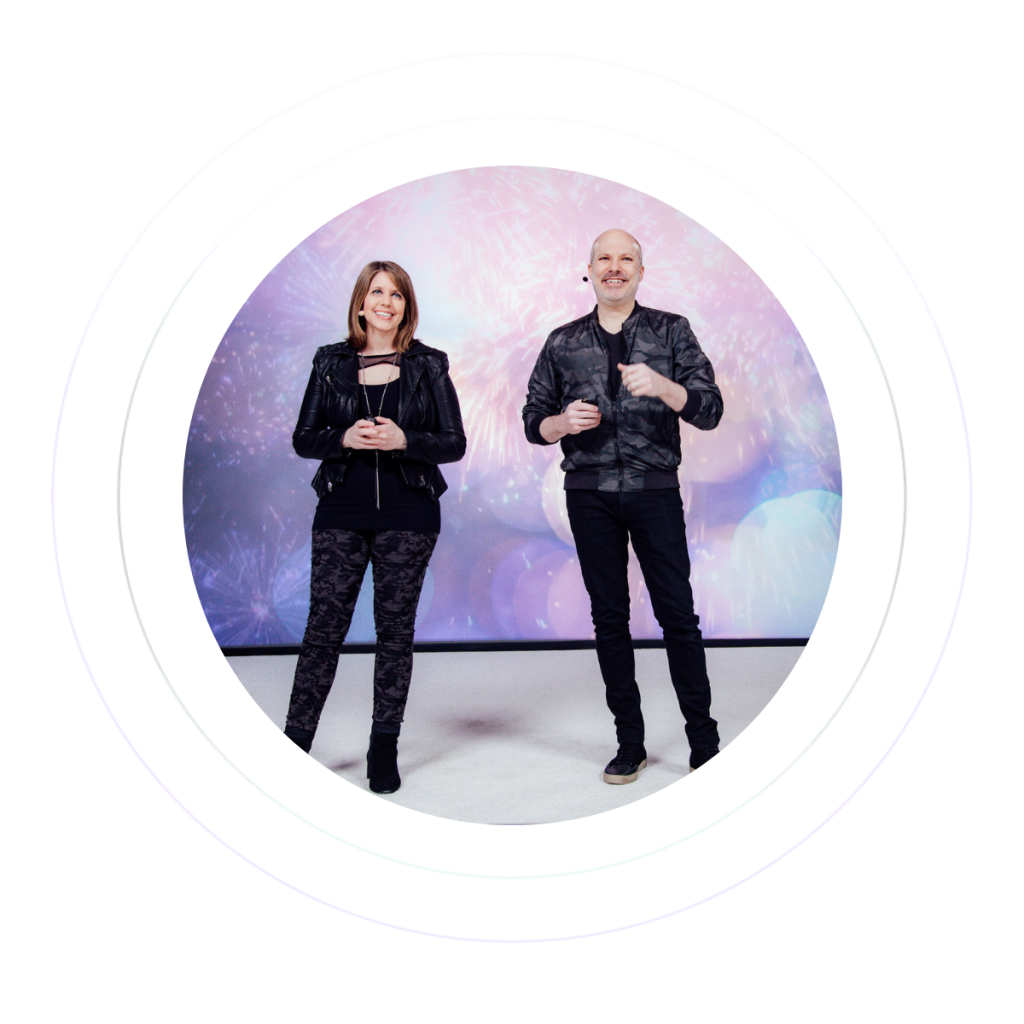 Steve and Jaime Geffner are wearing matching black outfits while speaking on a stage with fireworks in the background.